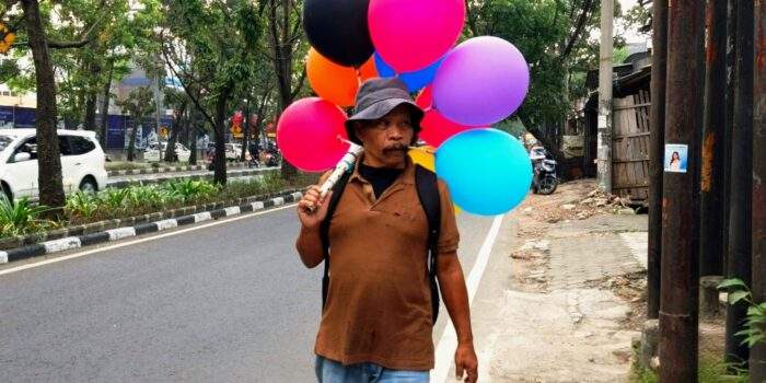 Seorang penjual balon keliling membawa puluhan balon warna-warni berjalan kaki menyusuri jalanan protokol di Kota Bandung pada sore hari.
